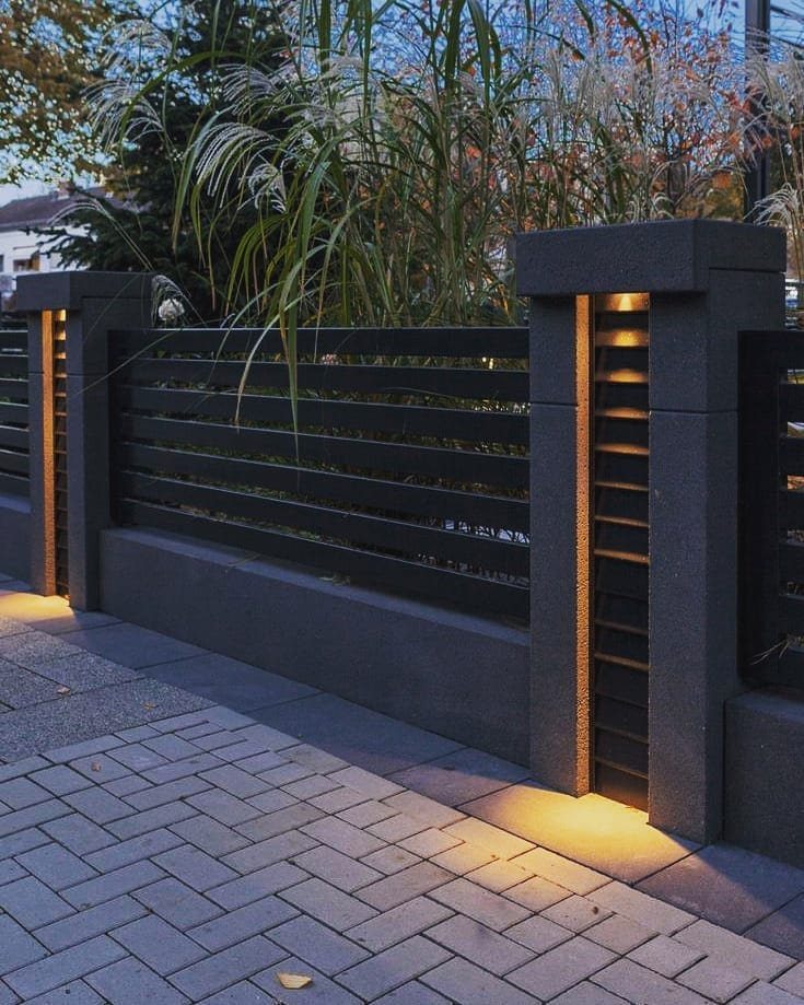 Modern dark grey fence with horizontal slats and illuminated pillars beside a brick path.