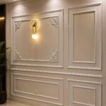 White decorative wall paneling with rectangular moldings and a gold wall sconce on a grey wall.