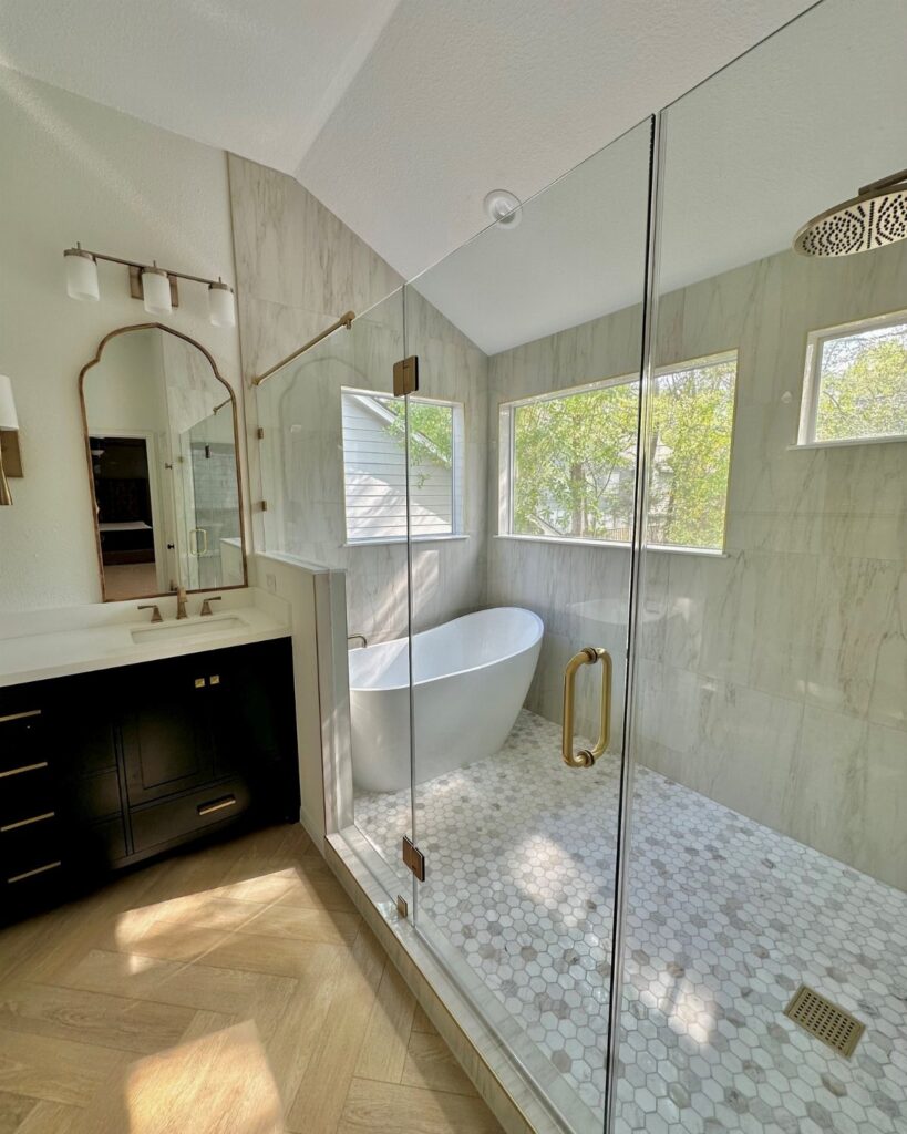 Modern bathroom with a glass shower, white bathtub, and light wood herringbone flooring.