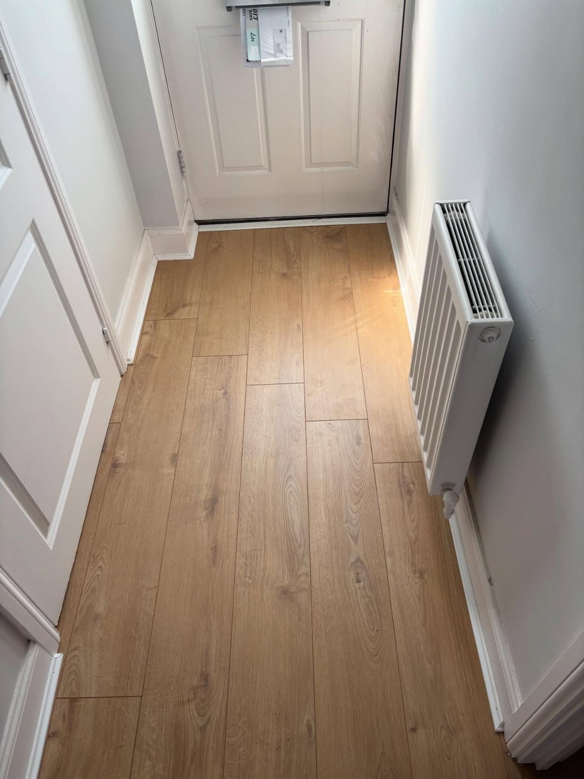 Light brown wood-effect laminate flooring planks in a narrow white hallway.